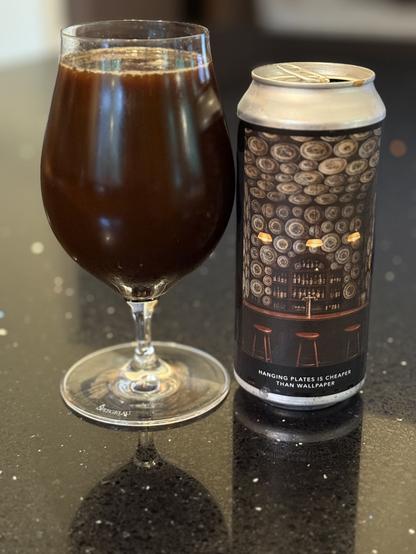 A tall snifter full of deep brown beer is next to a pint can showing a bar scene with walls and ceilings covered in patterned plates, and 3 bar stools along the rail.