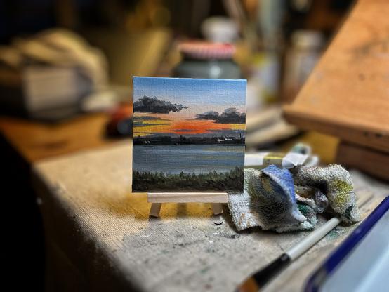 Alt text: A miniature painting on a tiny easel depicts a vivid sunset over water, with dark clouds above, orange and yellow hues at the horizon, and a dark green shoreline in the foreground. The small canvas sits on a cloth-covered work surface, surrounded by paintbrushes and a paint-stained rag, with a softly blurred artist’s studio in the background.