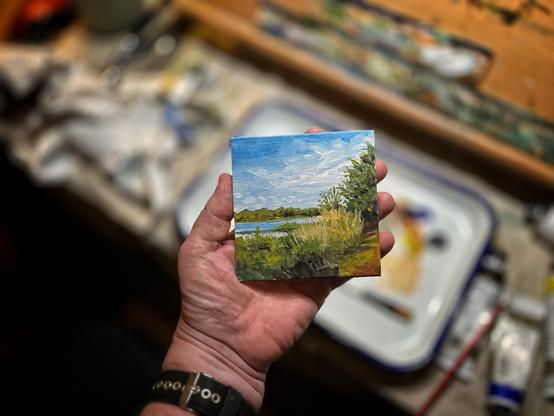 A hand holding a small square landscape painting depicting a lakeside scene with blue sky, water, green trees, and golden grasses, with art supplies and a palette visible in the blurred background.​​​​​​​​​​​​​​​​
