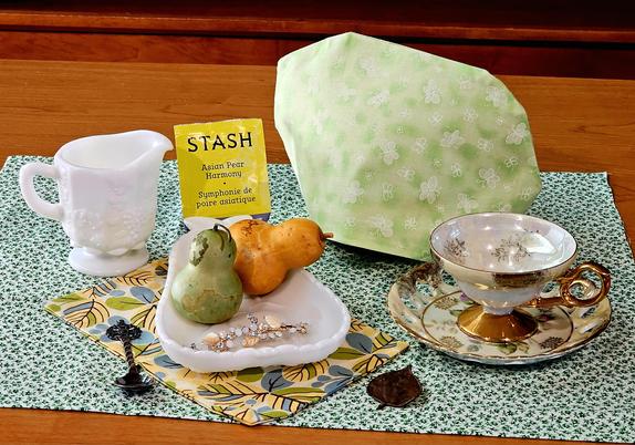 Tea tableau in spring green and warm yellow, with a pear- shaped Westmoreland milk glass dish holding a green Pear and a yellow pear and a jeweled brooch. There is a Westmoreland Paneled Grape milk glass cream pitcher, and an antique cup and saucer gleaming with gold leaf. An ornate bronze spoon and a bronze leaf rest near the Pear dish. The tea cozy and other linens provide a blend of cheerful florals, and the tea is Asian Pear white tea from Stash.