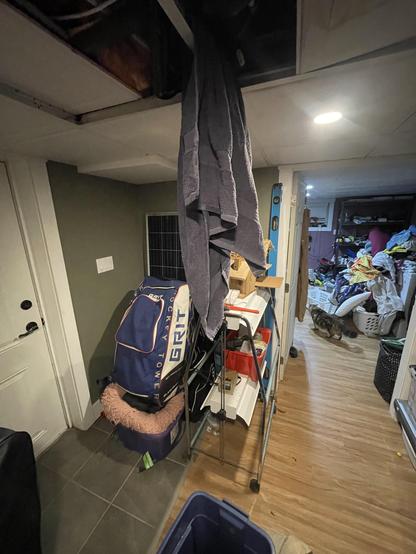 a blue towel hangs from the metal support of a drop tiled ceiling  as drips of water come down onto the floor of a finished basement.