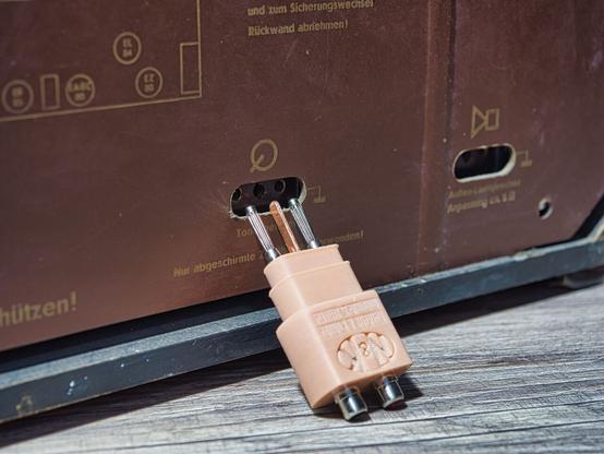 NF TA Stereo Stecker, Detailansicht, welche die Steckerlemente zeigt, an der Rückwand eines Röhrenradios.
Radioseitig sind 2 Bürstenstecker und eine Kupferschiene zu sehen. Auf der anderen Seite sind die beiden Cinch Anschlüsse.
Auf dem Stecker ist das N&K Logo zu erkennen.