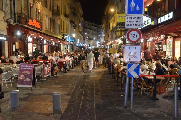 Photo of a pedestrian-only street in Nice, France.  The street is packed with people dining on patios and shopping and just enjoying themselves.  In the foreground are retractable bollards that block vehicle entrance at most times.  There is a sign saying in French that deliveries are allowed between 4am and 10:30am