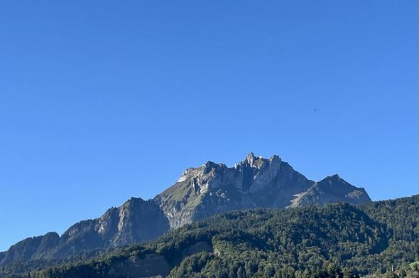 Pilatusberg, von der Sonne beschienen, vor wolkenlosem blauen Himmel. Die Bäume auf den bewaldeten Hügeln vor dem Berg zeigen vereinzelt schon einen Hauch Herbstfarben