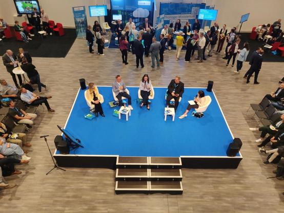 Blick von oben auf eine Bühne, auf der Bühne sitzen Milen Starke (Thüringer Digitalministerium), Stephan König (Staatskanzlei Thüringen), Esther Steverding (Bitkom), Timo König (Open Source Business Alliance) und die Moderatorin Blanka Weber. Um die Bühne herum sitzen Zuhörende auf Stühlen, hinter der Bühne sind außerdem Messestände zu sehen und eine Vielzahl von Personen, die sich an den Ständen austauschen.