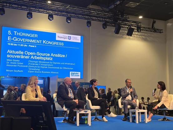 Auf einer Bühne sitzen Milen Starke (Thüringer Digitalministerium), Alexander Pockrandt (Zentrum für digitale Souveränität), Miriam Seyffarth (Open Source Business Alliance), André Göbel (FITKO) und die Moderatorin Blanka Weber. André Göbel spricht gerade in ein Mikrofon, die anderen Personen sind ihm aufmerksam zugewandt. Hinter der Bühne steht auf einem großen Bildschirm das Thema des Panels: „Aktuelle Open-Source Ansätze / Souveräner Arbeitsplatz“ und die Namen der Diskussionsteilnehmenden. Hinter der Bühne sind außerdem Messestände zu sehen und eine Vielzahl von Personen, die sich an den Ständen austauschen.