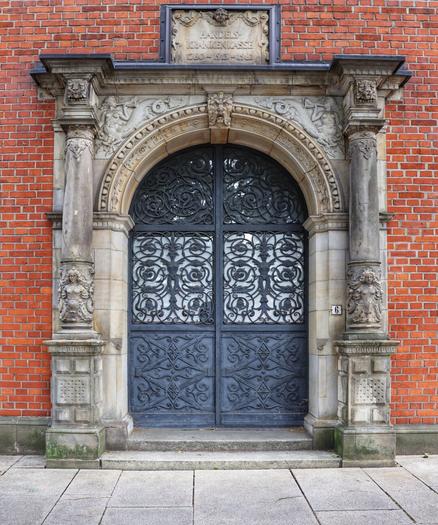 A richly detailed Renaissance-style stone portal with a dark grey, ornate wrought-iron double door set into a red brick wall. Above the arched doorway, an inscription reads "Handelskrankenkasse 1760–1913–1982". Two fluted columns with intricate capitals frame the arch, featuring grotesque masks and swirling foliage carvings. At the base of each column are sculptural figures. The number '6' is subtly visible to the right of the door.