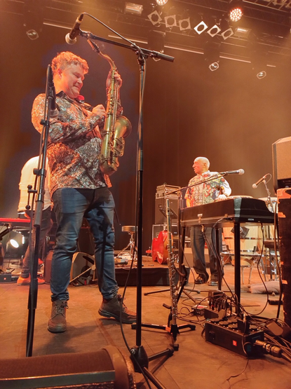 Einde van het concert. De saxofoonspeler lacht terwijl hij zijn instrument ontkoppelt. Mulatu Astatke staat op de achtergrond achter zijn elektrische piano en kijkt tevreden naar de pianist die net achter de saxofoonspeler over het podium loopt.