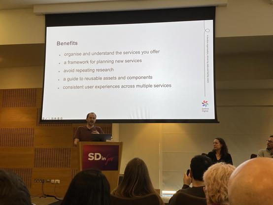 A middle aged white man behind a lectern in front of an audience with a projected slide above him saying: “Benefits 

—organise & understand the services you offer
—a framework for planning new services
—avoid repeating research
—a guide to reusable assets & components
—consistent user experiences across multiple services”