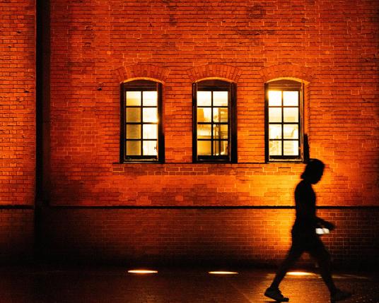 Brick wall with three arched windows, warmly lit from inside. Silhouette of a person walking in front, holding a phone, creating a contrast against the illuminated background.