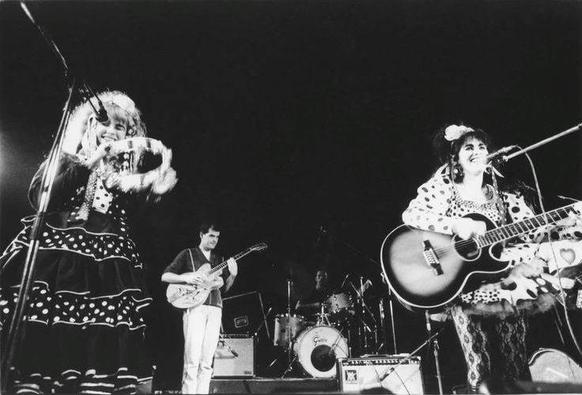 Black and white photo looking up at the stage. Jill Bryson is at the front left, smiling and playing a tambourine. Rose McDowall is at the front right, smiling and playing an acoustic guitar. In the centre further back is a drum kit on a riser played by Simon Dodds, in front of which stands Simon Emmerson playing guitar.