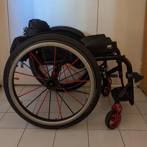 Photo d'un fauteuil roulant, de côté. Son dossier est bas. Il est noir avec un peu de rouge.