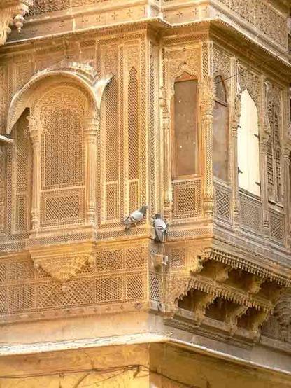 C'est une photos de l'un des angles de la façade d'une haveli . Taillés dans la pierre dorée, des moucharabiehs aux motifs multiples servent à la fois de fenêtres et de rafraîchisseurs en permettant à l'air de circuler. Leurs sculptures sont d'une grande finesse et font du bâtiment une oeuvre d'art. Au centre de l'image, deux pigeons semblent converser.