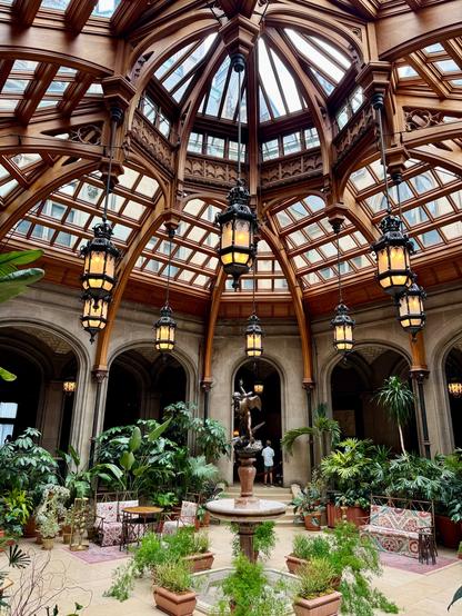 A round indoor room with a pergola-like domed, glass ceiling that lets in a lot of light for the garden setting full of tropical plants, palms and ferns. It has a fountain in the center and seating around the fountain. There is also a small round table at one seating arrangement.  The ceiling is all wood and glass, and there eight hanging lanterns made of wrought iron and opaque amber glass. This is the indoor conservatory room in the Biltmore House, Asheville, North Carolina.
