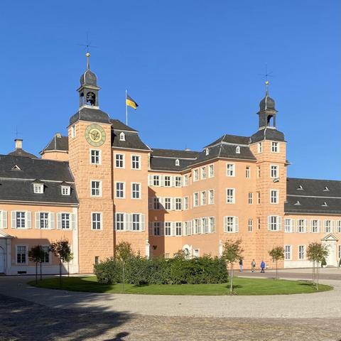 Das Bild zeigt ein barockes Gebäude mit einer pastellrosa Fassade und einem dunkelgrauen Dach. Es hat mehrere Stockwerke und ist durch zwei markante Türme mit Zinnen und Uhren gekennzeichnet. Die Türme sind mit goldenen Spitzen und Weckersternen verziert. Auf dem Dach weht eine Flagge mit den Farben Schwarz und Gold. Das Gebäude ist umgeben von einem gepflegten Rasen und Bäumen, und es gibt eine kleine Gruppe von Menschen, die auf dem Rasen spazieren. Der Himmel ist klar und blau.