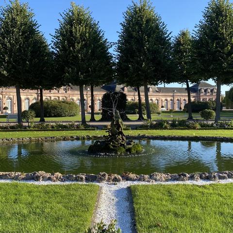 Das Bild zeigt einen barocken Garten mit einem zentralen Teich, der von einem kunstvollen Brunnen mit einer Statue in der Mitte umgeben ist. Der Teich ist von einem Rand aus Steinen und weißen Kieseln umgeben, der den Pfad zur Mitte des Teiches markiert. Im Hintergrund erhebt sich ein barockes Gebäude mit vielen Fenstern und einer klassischen Architektur, das von gepflegten Rasenflächen und Bäumen umgeben ist. Die Bäume sind symmetrisch angeordnet und tragen dichte, grüne Blätter. Der Himmel ist klar und blau.