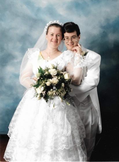 Wedding photo of a young woman with her honey-blonde hair pulled back and in a light lacy veil. Her wide-skirted white gown is very lace-themed. Behind her and to her side is a young man in a white suit, with his arms wrapped around her from behind. He has jet-black hair and glasses with large lenses. She is reaching back with one hand to rest her finger on his cheek. In front of them both is a bouquet of white flowers.