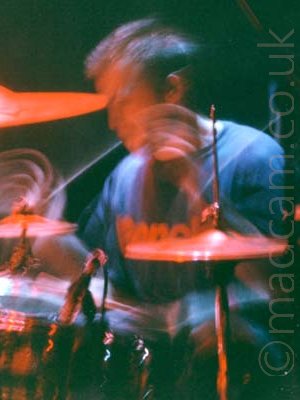 A man with short dark hair, wearing a dark blue t-shirt with indistinct red text across the chest, sitting behind a black and chrome drum kit. A long exposure and his movement has streaks of light as his drumsticks move, lit red by the stagelights, along with the rest of the kit and his face. Black walls fill the background.