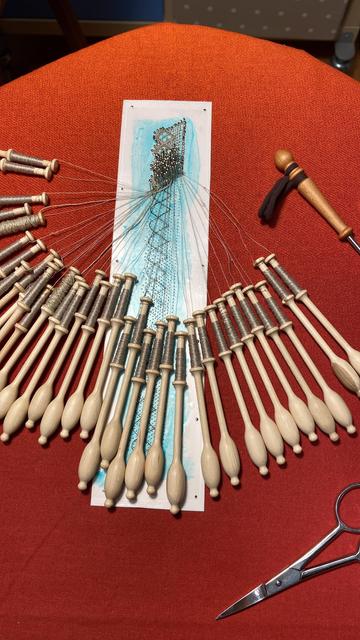 Ein Flachkissen, auf dem ein Pappstreifen mit vielen Nadeln steckt, daran 36 Klöppel oder 18 Paar mit dünnem Garn