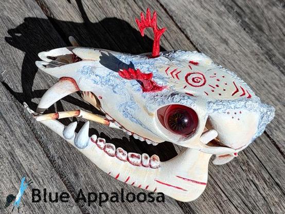 Top/side view of a skull of a deer-like creature with prominent fangs on the upper jaw, a split lower-with a a pair of fangs, and antennae-like structures protruding from the mouth. The skull is tan-colored, with parts coated in a light blue-ish substance. It also has red eyeballs and red, feathery antennae coming from the top of it's head. There are red lines carved into the lower jaw. 


Watermark Reads: Blue Appaloosa