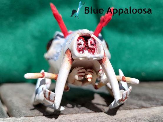 Front view of a skull of a deer-like creature with prominent fangs on the upper jaw, a split lower-with a a pair of fangs, and antennae-like structures protruding from the mouth. There is also a structure looking like a pair of hands with interlaced fingers. The skull is tan-colored, with parts coated in a light blue-ish substance. It also has a red-colored substance inside the nasal cavity and red, feathery antennae coming from the top of it's head. 


Watermark Reads: Blue Appaloosa