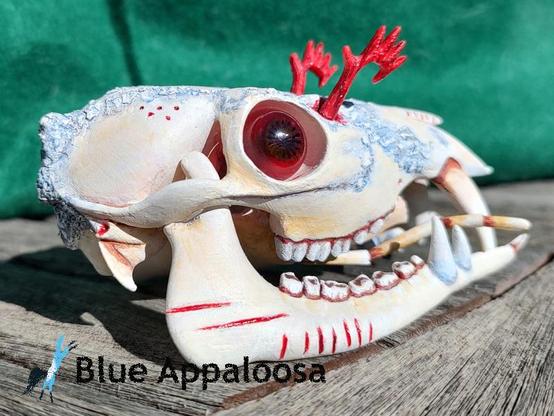 Back/side view of a skull of a deer-like creature with prominent fangs on the upper jaw, a split lower-with a a pair of fangs, and antennae-like structures protruding from the mouth. The skull is tan-colored, with parts coated in a light blue-ish substance. It also has red eyeballs and red, feathery antennae coming from the top of it's head. There are red lines carved into the lower jaw. 


Watermark Reads: Blue Appaloosa