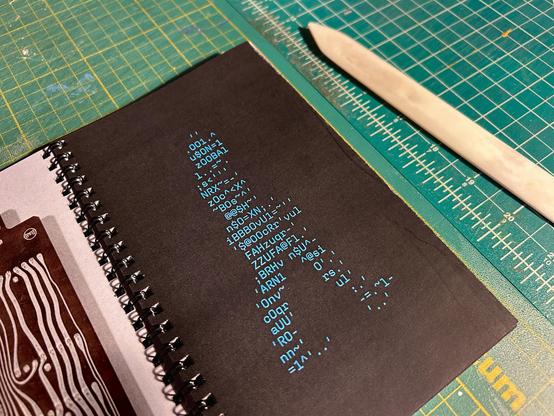 Pages from an open wire-bound book showing a walking figure made out of blue-colored ASCII characters on a black field opposite a vintage circuit board. In the background is a green cutting mat and a cream colored bone folder.