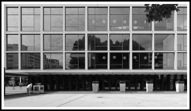 Das Schwarz-Weiß-Foto zeigt die Fassade eines modernen Gebäudes. Die Architektur ist geprägt von klaren Linien und geometrischen Formen. Die Fassade besteht aus einer regelmäßigen Anordnung großer Fensterflächen, die in einem Raster angeordnet sind. Eine Reihe von gleichförmigen, rechteckigen Pflanzgefäßen steht vor dem Gebäude. An der linken Ecke ist ein Mann zu sehen, der sich im Bildausschnitt sehr klein darstellt, was einen Kontrast zwischen der Größe des Gebäudes und der Person erzeugt. Der Mann betrachtet heruntergebeugt die Inschrift auf dem Sockel einer kleinen Skulptur.

The black-and-white photograph shows the facade of a modern building. The architecture is characterized by clean lines and geometric shapes. The facade consists of a regular arrangement of large windows arranged in a grid pattern. A row of uniform, rectangular planters stands in front of the building. In the left corner, a man can be seen, who appears very small in the frame, creating a contrast between the size of the building and the person. The man is bent over, looking at the inscription on the base of a small sculpture.