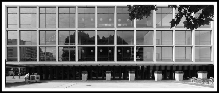 Das Schwarz-Weiß-Foto zeigt die Fassade eines modernen Gebäudes. Die Architektur ist geprägt von klaren Linien und geometrischen Formen. Die Fassade besteht aus einer regelmäßigen Anordnung großer Fensterflächen, die in einem Raster angeordnet sind. Eine Reihe von gleichförmigen, rechteckigen Pflanzgefäßen steht vor dem Gebäude. An der linken Ecke ist ein Mann zu sehen, der sich im Bildausschnitt sehr klein darstellt, was einen Kontrast zwischen der Größe des Gebäudes und der Person erzeugt. Der Mann betrachtet heruntergebeugt die Inschrift auf dem Sockel einer kleinen Skulptur.

The black-and-white photograph shows the facade of a modern building. The architecture is characterized by clean lines and geometric shapes. The facade consists of a regular arrangement of large windows arranged in a grid pattern. A row of uniform, rectangular planters stands in front of the building. In the left corner, a man can be seen, who appears very small in the frame, creating a contrast between the size of the building and the person. The man is bent over, looking at the inscription on the base of a small sculpture.