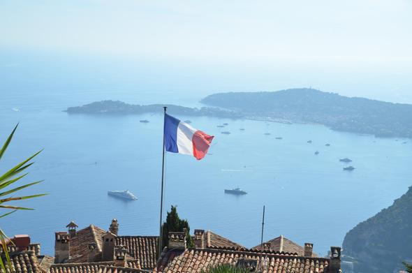 Photo of the view from high up a mountain.  In the foreground are the roofs of medieval homes, one flying a French flag.  In the background is the sea far below, and an island.  In the water are numerous large yachts.