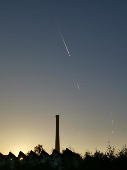 Skyline d'anciens bâtiments industriels, et une grande cheminée, sur un ciel de soleil levant sans nuages marqué de 3 ou 4 traces d'avions.