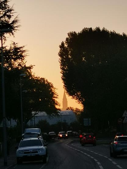 Dans l'axe d'une rue sinueuse, où des automobiles circulent, feux allumés, au jour levant. Sur les côtés, des arbres, à contre jour, et au fond, dans une trouée, un clocher nimbé dans un ciel orange.