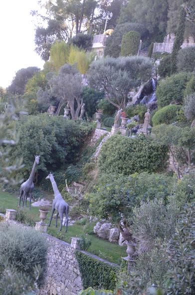 Photo of two terraces of a garden carved into the side of a mountain.  On the upper terrace lawn are several large Greek urns, and life/sized statues of a lion, some women, a horse and a pony.  On the lawn of the lower terrace are life-sized statues of two giraffes.  There is a stone retaining wall below each terrace.