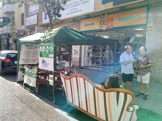Una paradeta al carrer amb carpa verda, un sofà, una taula, cadires, cartells de la cooperativa Ecotxe i algunes persones conversant.