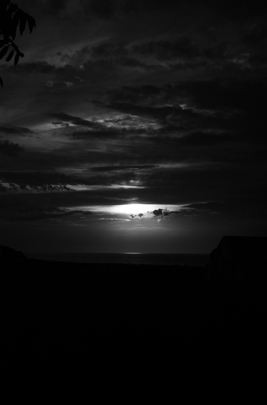 A black and white photography depicting a dark sky with a view on the sea. The sun is just behind a cloud.