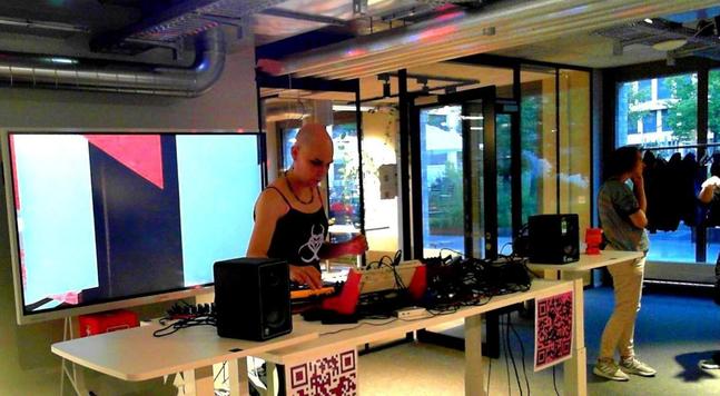 CyberGwen in concert, standing behind a standing desk full of synths and other gear, with a visualizer on a big screen behind her.
Photo by seism CC BY 4.0