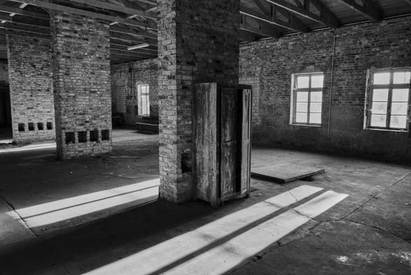 Blick in einen großen, leeren Raum. In dem Raum stehen mehrere ehemalige Schornsteinzüge. Der Raum wurde früher als Großküche genutzt.  Im Hintergrund befinden sich mehrere Fenster. Durch sie fällt Sonnenlicht schräg in den Raum hinein.