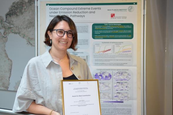 Danai Filippou is standing in front of her poster "Ocean Compound Extreme Events under Emission Reduction and Negative CO2 pathways" and holding a frame with a certificate saying "Award for Best Contribution"