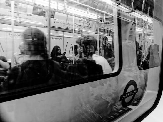 Black and white photograph of passengers inside a London Underground train, seen through the reflective window, blending sharp details with ghostly overlays of reflections.