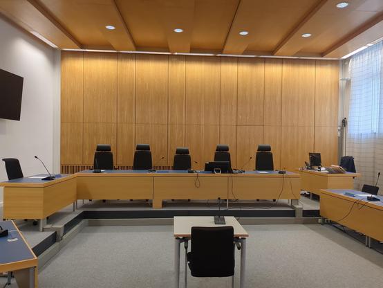 Blick auf einen menschenleeren Gerichtssaal, auf die Richterbank. Graue getäfelte Rückwand. Rechts Vorhänge in weiß, links die Bank der Staatsanwaltschaft
