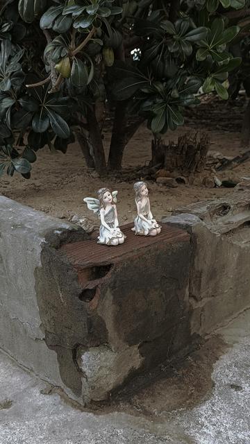 The piss-stained corner of a pavement's raised bed. The plaster is broken enough to reveal a brick underneath. Someone has neatly placed two small, light-coloured fairies on it. Probably plastic, painted to imitate porcelain. 
They are both sitting with their feet tucked under, holding flowers and smiling calmly. The left one has little wings on her back.
They seem to ignore the dirt they are sitting on.
Behind them a few dusty pittosporum bushes, the kind we call "Angelica" here.