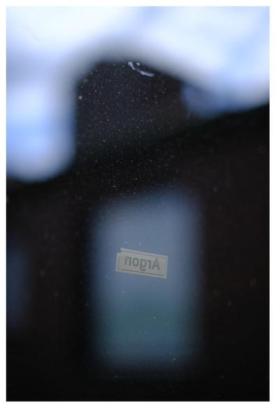Blick durch ein staubiges Fenster mit Aufkleber "Argon" auf ein dunkles Gebäude, an dem man ein etwas helleres Fenster ahnt. Das Gebäude hat einen kleinen Turm. Das Bild ist recht dunkel.