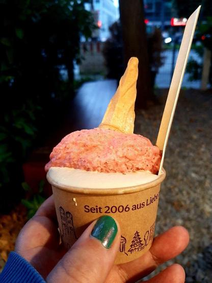 Ein Pappbecher mit Eis udn einer Waffel und einem Waffel drin, er wird von meiner Hand ins Bild gehalten, im Hintergrund Straßen und Büsche bei ziemlicher Dunkelheit.