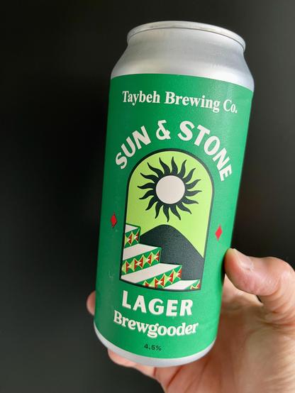 a hagard hand (mine) grasps a beautiful, 440ml can of sun & stone lager, which is a collaboration between taybeh brewing company in palestine & brewgooder brewing in scotland. 
the tinnie is predominantly shades of green. under the name is an arched window shape, through which one can see steps decorated with green & red tiles. behind the steps is the suggestion of a mountain & above the mountain is a full sun with stylised rays.