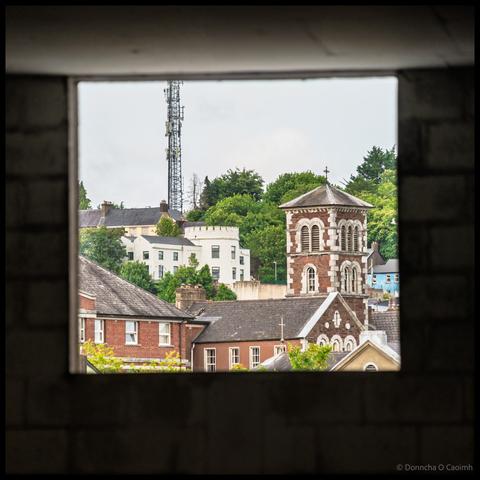 View through concrete window frame of Cork cityscape showing red brick Victorian church with ornate tower, mixed residential buildings, telecommunications mast, and lush green trees from elevated perspective of North Main Street multi-storey car park.