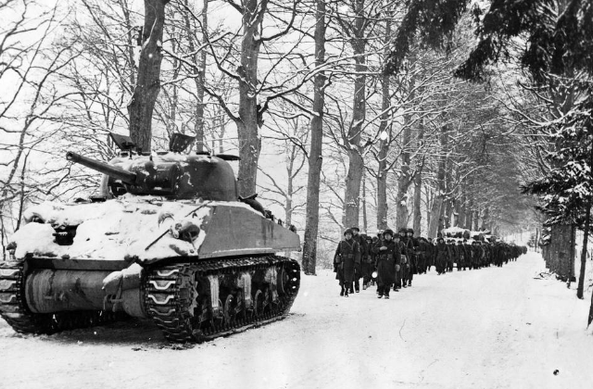 A photo of the Battle of the Bulge. A snowy tank is leading a long line of soldiers, all marching down a snow covered road lined with trees.