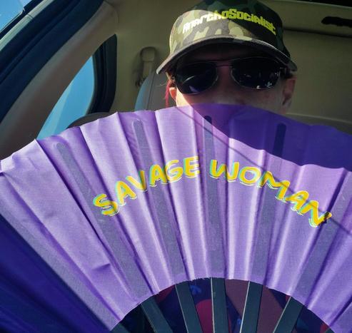 Person wearing sunglasses and an "AnarchoSocialist" hat inside a car, holding a purple fan with "SAVAGE WOMAN" printed on it in yellow and turquoise.