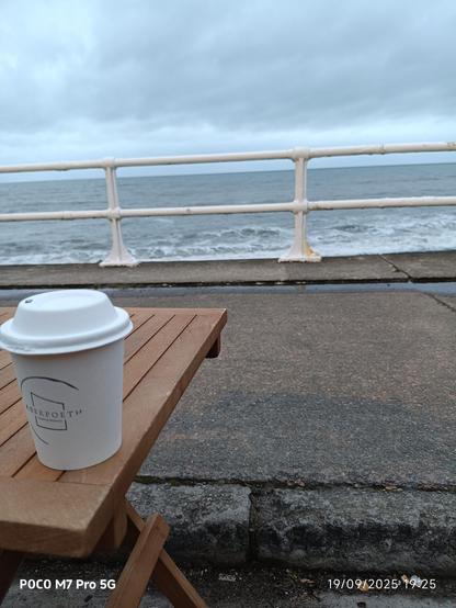 View of the sea through stone railings, the horizon just below the top rail. 

In the foreground is a small wooden table with a coffee cup on the corner.

The sky is grey cloud.