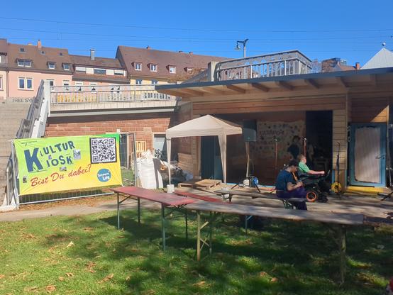 Sicht auf den aus Holz im Bau befindlichen Kultur Kiosk.
Links ein Treppenaufgang (zur Brücke). Darunter ein Banner: " Kulturkiosk - Bist Du dabei" in hellen bunten Farben.

 Einige leere Biertische und Bierbänke stehen auf dem Rasen. Eine Person im Rollstuhl am Eingang des aus Holz gebauten Kiosk.

Im Hintergrund sieht man noch die Spitzen einiger Hausdächer.