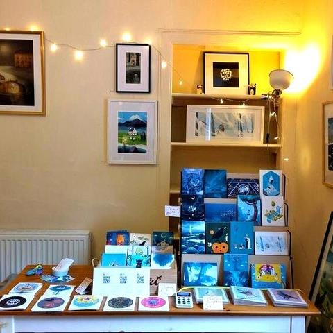 Photo of a table with lots of postcards, stickers and greeting cards with my art on it, in the background there are framed prints and fairy lights.